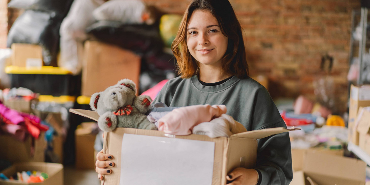 Eine Jugendliche hält einen Karton mit einem Teddybären und Kleidung. Im Hintergrund weitere Kartons mit Sachspenden.