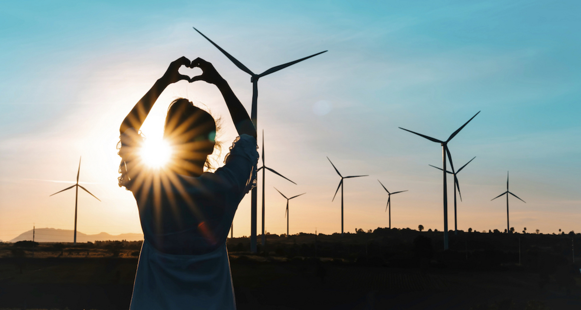 Ein Mensch steht im Freien vor einer Reihe von Windrädern und formt mit den Händen ein Herzsymbol. Der Sonnenuntergang erzeugt einen warmen Schein, der die Silhouette der Person und der Windräder kontrastreich hervorhebt. Der Himmel ist klar und blau, und die Szene vermittelt eine Botschaft von Wertschätzung für erneuerbare Energien und die Natur.