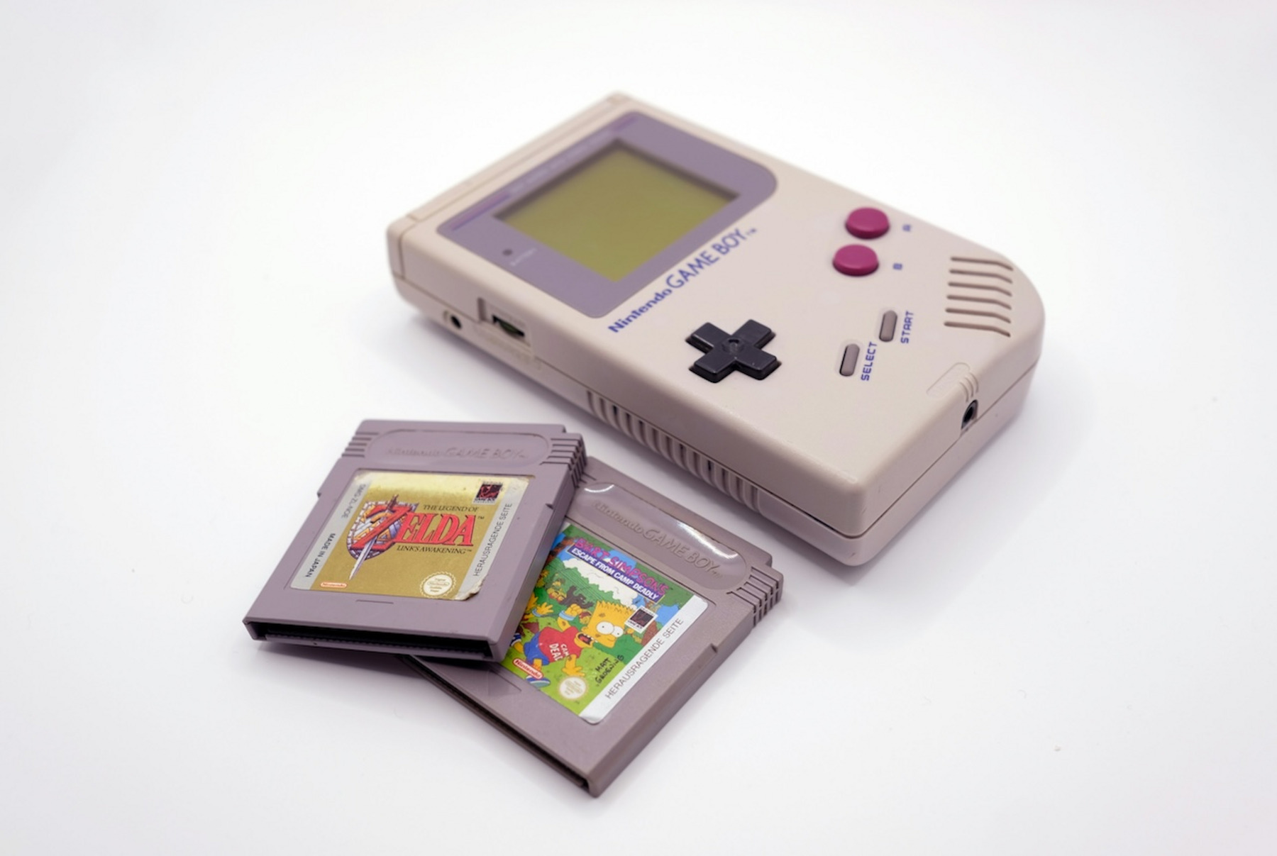 Ein Nintendo Game Boy mit zwei Spielemodulen, eines davon „The Legend of Zelda: Link‘s Awakening“ und das andere „Bart Simpson‘s Escape from Camp Deadly“. Der Game Boy ist beige mit grauen und lila Tasten.