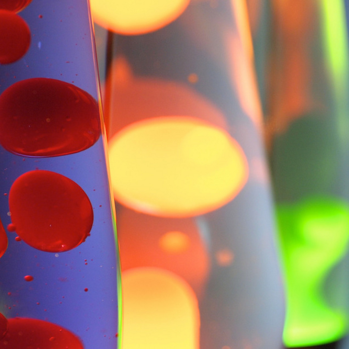 Eine Nahaufnahme einer Lavalampe mit leuchtenden, roten Blasen, die in einer klaren Flüssigkeit schweben. Im Hintergrund sind verschwommene Lichter in Gelb und Grün von weiteren Lavalampen zu sehen.