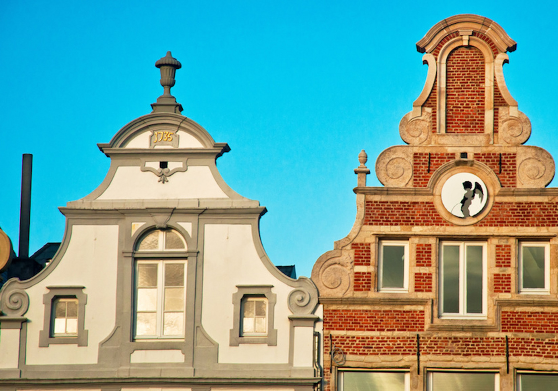 Zwei Giebelhäuser mit dekorativen Fassaden am Großen Markt in Mechelen, eines in Weiß und das andere in Rotstein, im typisch flämischen Baustil. Beide Fassaden haben geschwungene Giebel und historische Details.