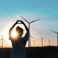 Ein Mensch steht im Freien vor einer Reihe von Windrädern und formt mit den Händen ein Herzsymbol. Der Sonnenuntergang erzeugt einen warmen Schein, der die Silhouette der Person und der Windräder kontrastreich hervorhebt. Der Himmel ist klar und blau, und die Szene vermittelt eine Botschaft von Wertschätzung für erneuerbare Energien und die Natur.