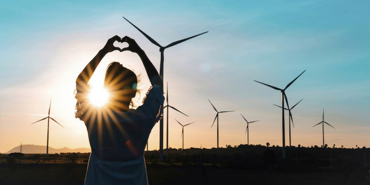 Ein Mensch steht im Freien vor einer Reihe von Windrädern und formt mit den Händen ein Herzsymbol. Der Sonnenuntergang erzeugt einen warmen Schein, der die Silhouette der Person und der Windräder kontrastreich hervorhebt. Der Himmel ist klar und blau, und die Szene vermittelt eine Botschaft von Wertschätzung für erneuerbare Energien und die Natur.