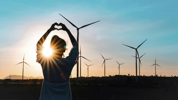 Ein Mensch steht im Freien vor einer Reihe von Windrädern und formt mit den Händen ein Herzsymbol. Der Sonnenuntergang erzeugt einen warmen Schein, der die Silhouette der Person und der Windräder kontrastreich hervorhebt. Der Himmel ist klar und blau, und die Szene vermittelt eine Botschaft von Wertschätzung für erneuerbare Energien und die Natur.