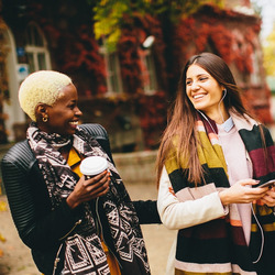 Zwei junge Frauen schlendern durch eine herbstliche Stadt und lachen miteinander.