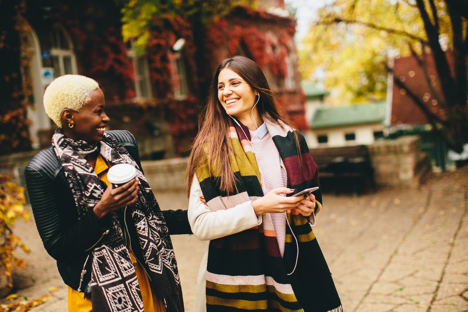 Zwei junge Frauen schlendern durch eine herbstliche Stadt und lachen miteinander.