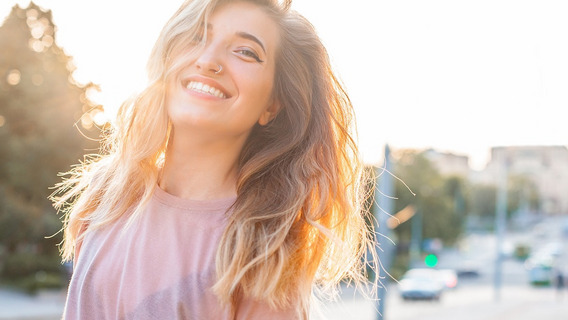 Eine blonde Frau steht lachend auf einer Straße in der Sonne. Im  Hintergrund sind Menschen, Autos und Bäume zu sehen.