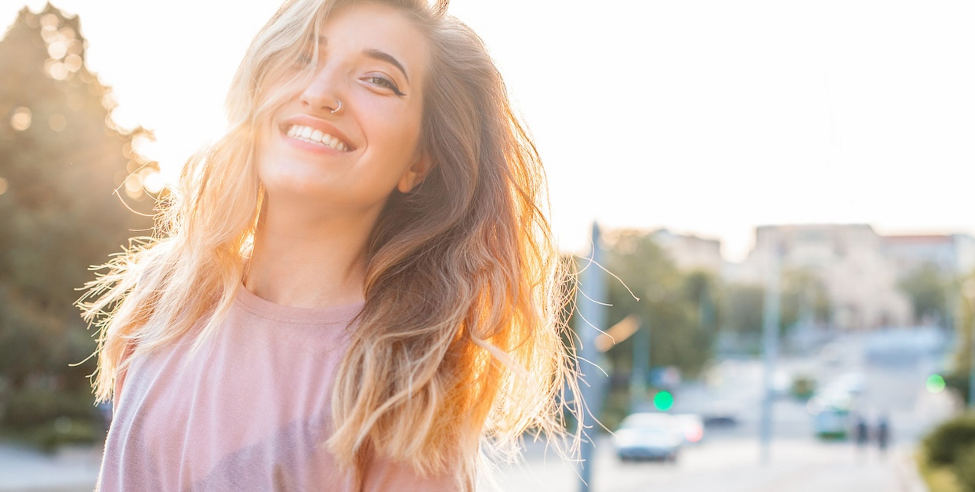 Eine blonde Frau steht lachend auf einer Straße in der Sonne. Im  Hintergrund sind Menschen, Autos und Bäume zu sehen.