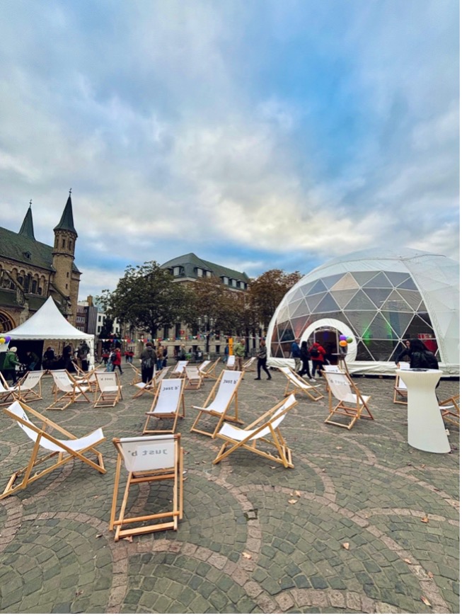 Ein belebter Stadtplatz mit historischen Gebäuden im Hintergrund. Im Vordergrund stehen mehrere Liegestühle aus Holz. In der Mitte des Platzes befindet sich eine große, weiße geodätische Kuppel. Menschen laufen über den Platz und einige kleine Zelte sind aufgebaut. Der Himmel ist bewölkt mit blauen Durchbrüchen.