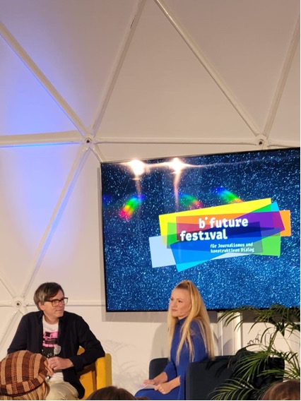 Zwei Personen sitzen auf einem Sofa vor einem großen Bildschirm. Links sitzt ein Mann mit Brille und dunklem Hemd, rechts eine Frau mit langen blonden Haaren. Der Bildschirm im Hintergrund zeigt ein buntes Logo mit der Aufschrift "b°future festival". Die Szene spielt sich in einem Raum mit gewölbter Decke ab.