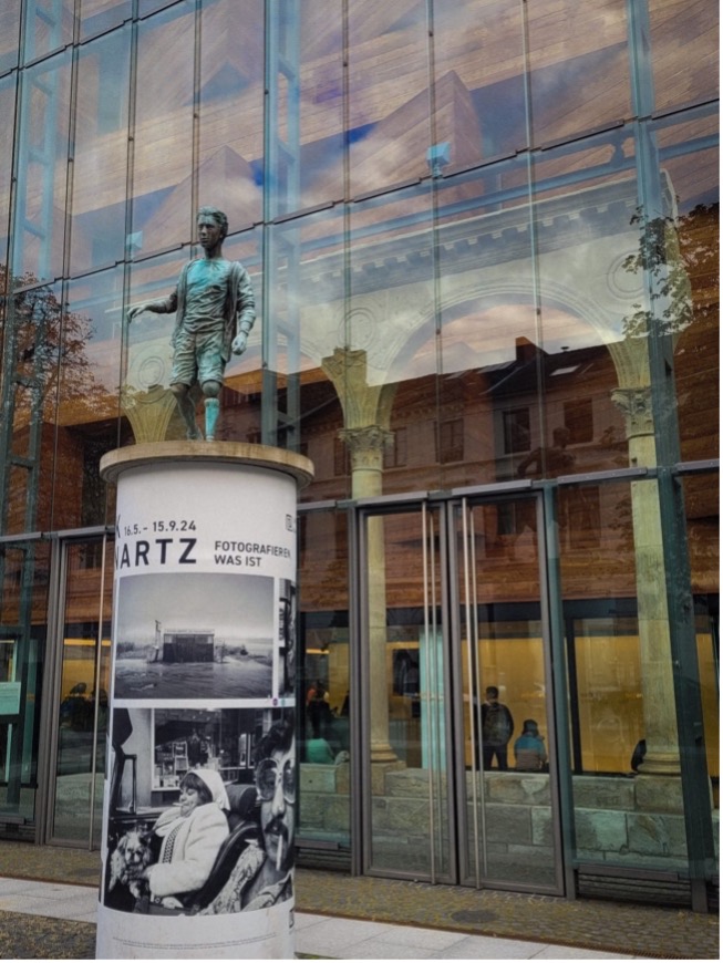 Ein modernes Glasgebäude spiegelt den Himmel und umliegende Gebäude wider. Im Vordergrund steht eine Säule mit Werbung für eine "ARTZ" Ausstellung, auf der eine Schwarzweißfotografie zu sehen ist. Auf der Säule befindet sich eine bronzefarbene Statue eines Menschen in aufrechter Pose. Das Gebäude hat große Glasfenster und eine reflektierende Fassade.